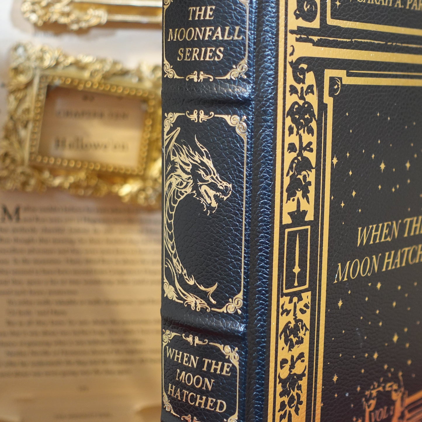 A close-up of a hardcover book titled 'When the Moon Hatched' from The Moonfall Series, featuring a dark navy blue cover with gold embossed details and a dragon illustration on the spine, set against a blurred vintage book background.