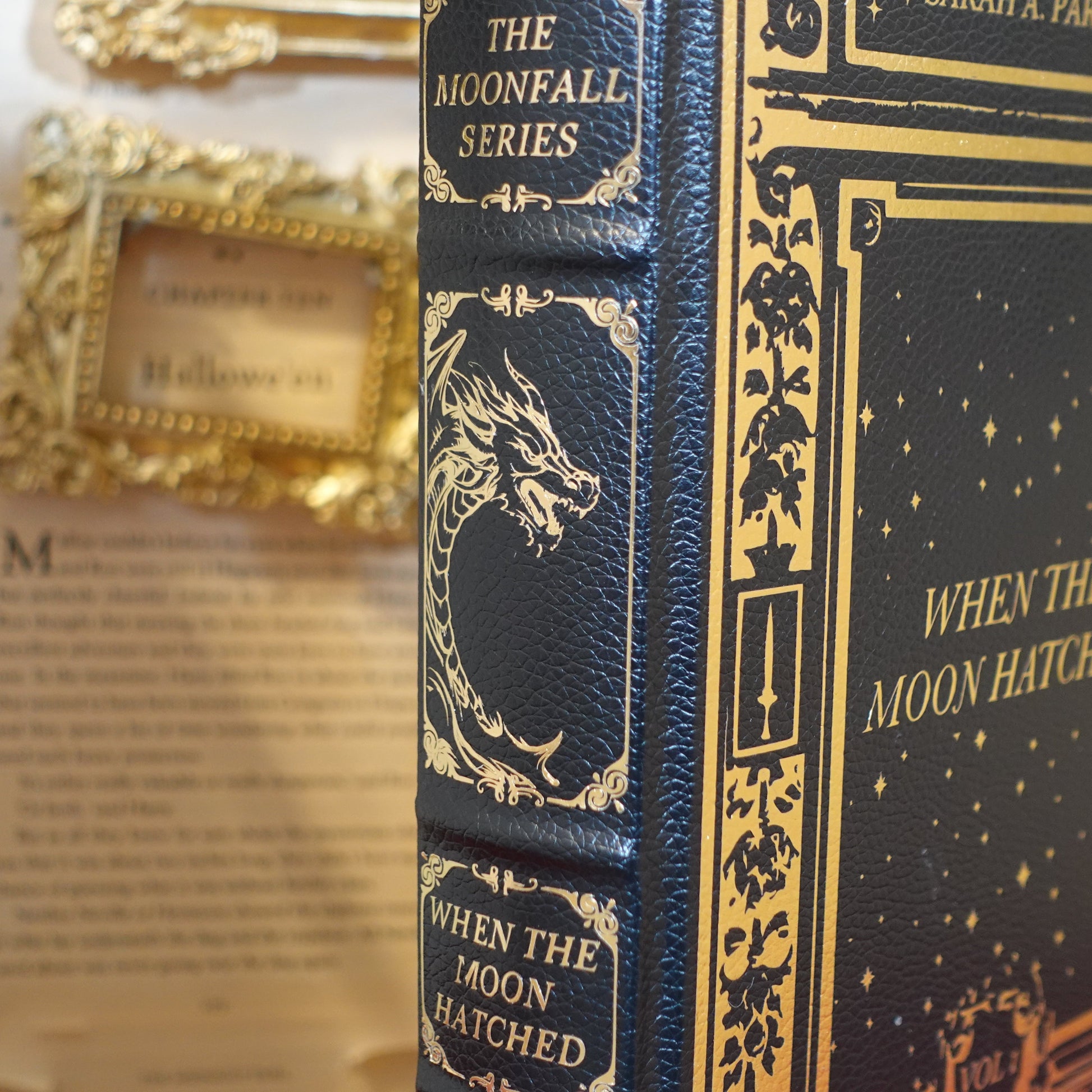 A close-up of a hardcover book titled 'When the Moon Hatched' from The Moonfall Series, featuring a dark navy blue cover with gold embossed details and a dragon illustration on the spine, set against a blurred vintage book background.