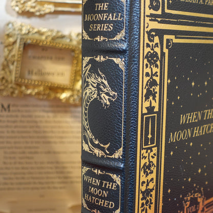 A close-up of a hardcover book titled 'When the Moon Hatched' from The Moonfall Series, featuring a dark navy blue cover with gold embossed details and a dragon illustration on the spine, set against a blurred vintage book background.